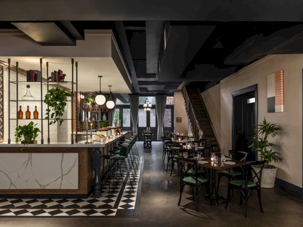 A stylish cafe with a marble bar, checkered floor, hanging lights, plants, and several seating tables along a dimly lit, modern interior.