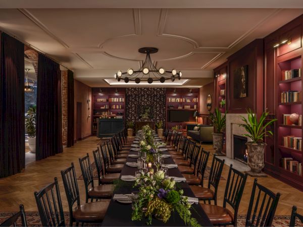 A long dining table with black chairs, floral centerpiece, warm wood shelves, books, plants, and soft chandelier lighting in an elegant room.