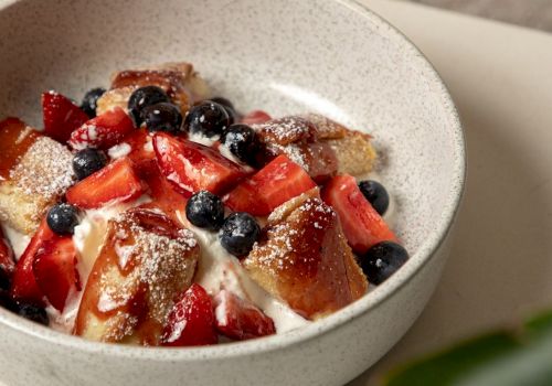 A bowl of dessert with cubed cake, whipped cream, strawberries, and blueberries topped with powdered sugar, in a light speckled bowl.