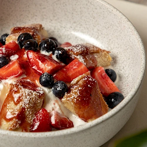 A bowl of dessert with cubed cake, whipped cream, strawberries, and blueberries topped with powdered sugar, in a light speckled bowl.