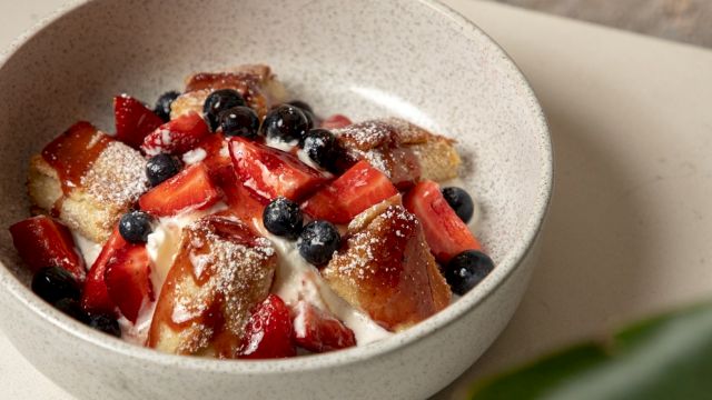 A bowl of dessert with cubed cake, whipped cream, strawberries, and blueberries topped with powdered sugar, in a light speckled bowl.