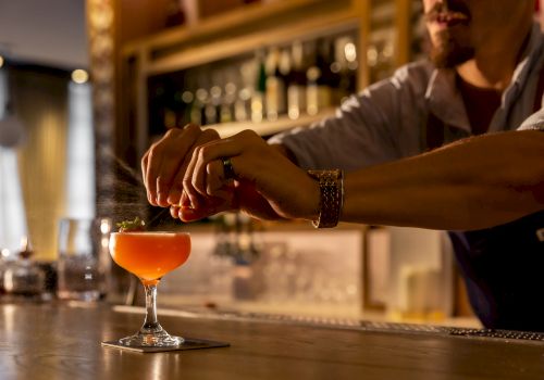 A bartender pours an orange cocktail at a dimly lit bar, with the drink in a coupe glass and a wristwatch visible on the arm. (ends with a period)
