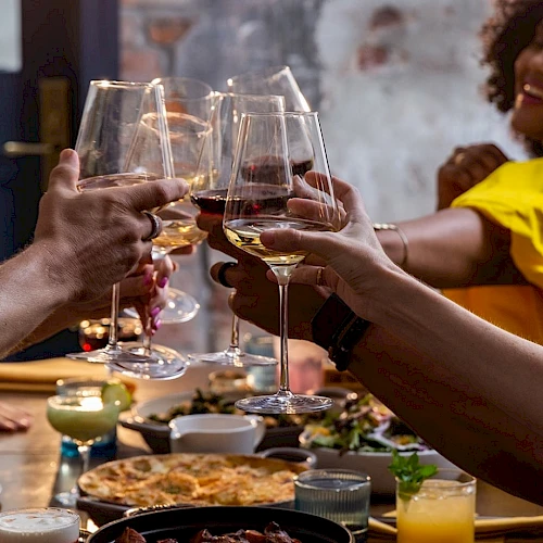 A group of friends raises glasses in a toast around a table full of shared dishes and drinks, celebrating together with smiles.