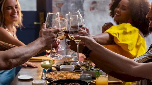 A group of friends raises glasses in a toast around a table full of shared dishes and drinks, celebrating together with smiles.