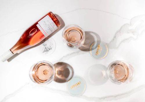 A pink ros&eacute; bottle pours into several small cups on a white marble surface, with caps and a few spilled droplets nearby.