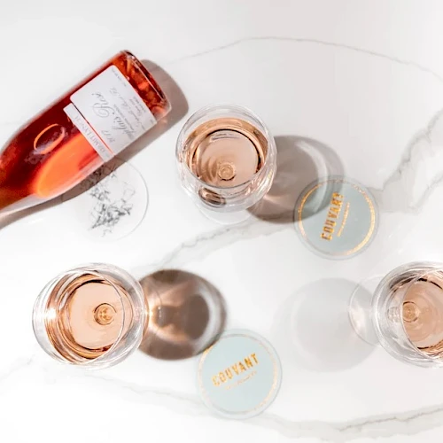 A pink ros&eacute; bottle pours into several small cups on a white marble surface, with caps and a few spilled droplets nearby.