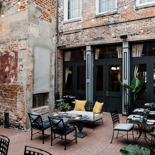 A stylish outdoor cafe in a brick-walled courtyard with black doors, mix of cushioned seating, metal chairs, and potted plants, warm and inviting.