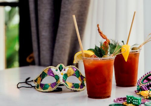 Two colorful Mardi Gras-style masks lie on a table beside two iced fruit drinks with lemon wedges and straws.