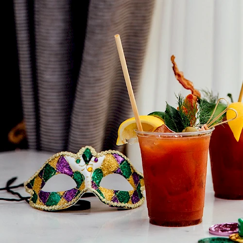 Two colorful Mardi Gras-style masks lie on a table beside two iced fruit drinks with lemon wedges and straws.