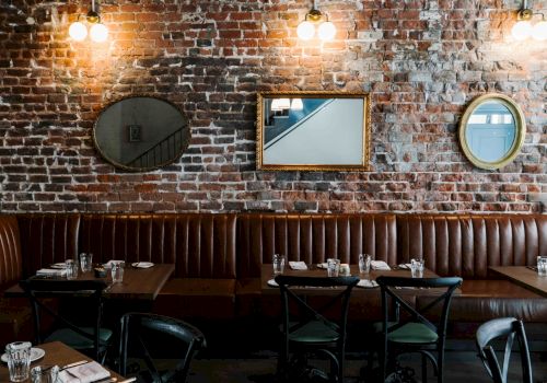 Cozy brick-walled restaurant with a long brown leather banquette, round mirrors, and warm hanging lights over dark wooden tables.