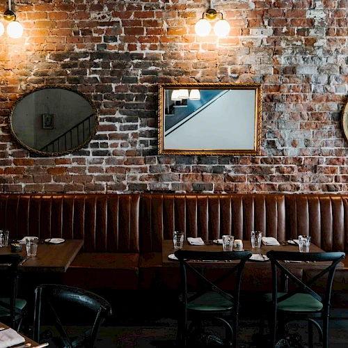 Cozy brick-walled restaurant with a long brown leather banquette, round mirrors, and warm hanging lights over dark wooden tables.
