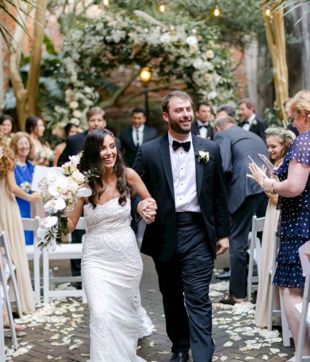 A newlywed couple walks down an aisle, holding hands, while guests applaud under a floral arch at their outdoor wedding.