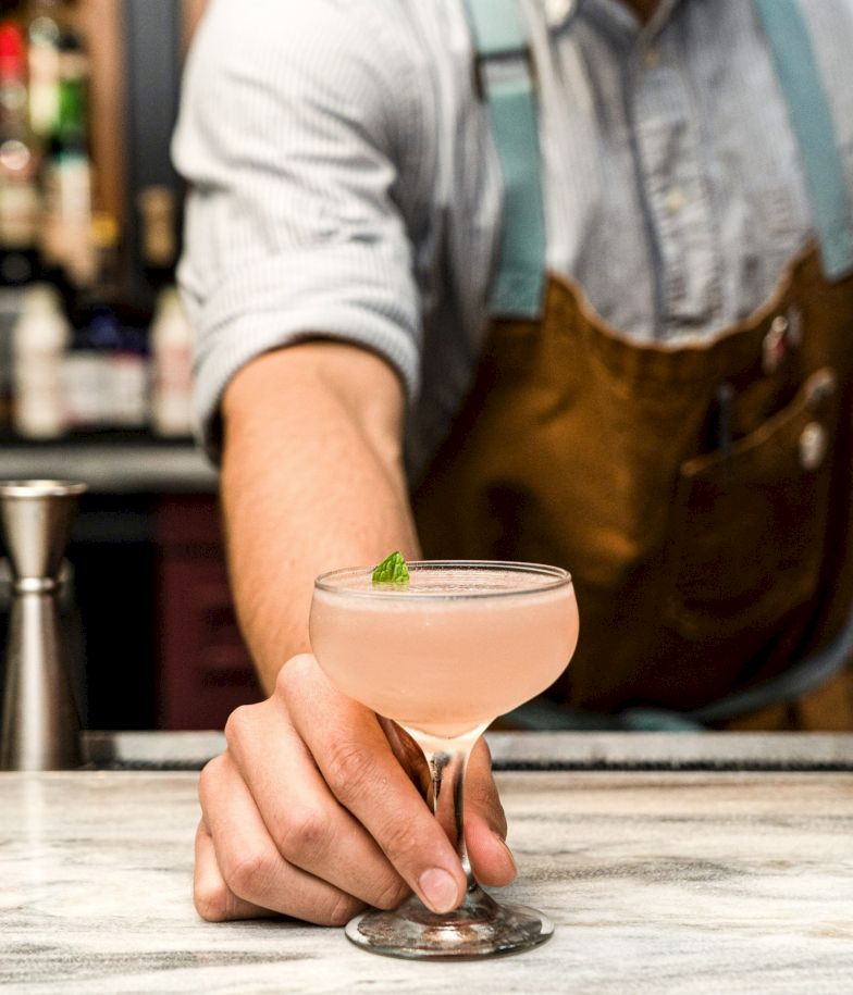 A bartender in a denim shirt and apron offers a pale pink cocktail with a lime garnish in a coupe glass at a marble bar, handheld gently.