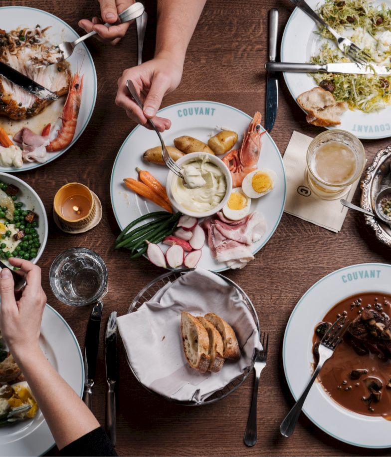 A table of varied dishes: roasted meat, veggies, bread, sauces, and drinks, with hands reaching in from all sides to share the feast.