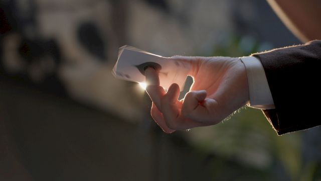 A hand holding a lit cigarette or joint, with a faint glow at the tip, in a suit sleeve, against a blurred background. End.