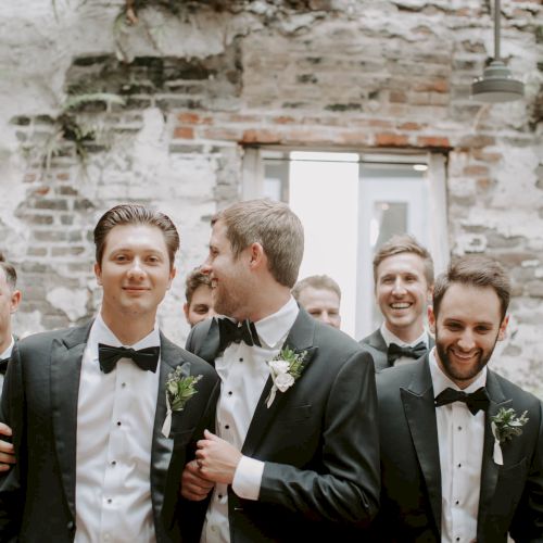 A group of men in tuxedos, likely groomsmen, smiling and toasting at a rustic outdoor wedding.