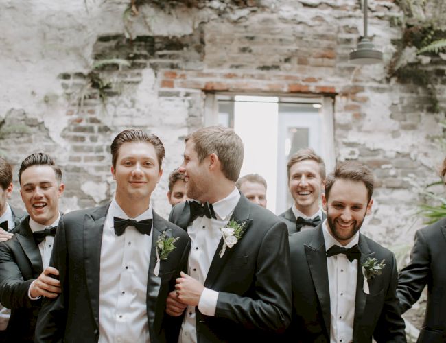 A group of men in tuxedos, likely groomsmen, smiling and toasting at a rustic outdoor wedding.