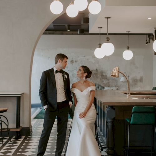 A newlywed couple smiles at each other in a stylish, modern venue with checkered floor, arc doorway, and chic hanging lights.