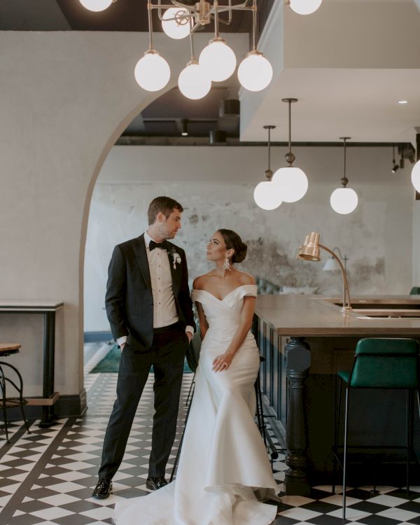 A newlywed couple smiles at each other in a stylish, modern venue with checkered floor, arc doorway, and chic hanging lights.