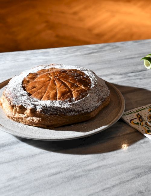 A round almond cake dusted with powdered sugar sits on a plate with a fork, while a pale cocktail with a lime wheel rests on a coaster beside it, on a marble table.