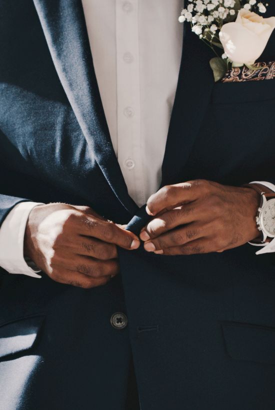 A man in a navy suit buttons his jacket, wearing a white shirt and a boutonniere; a wristwatch is visible.