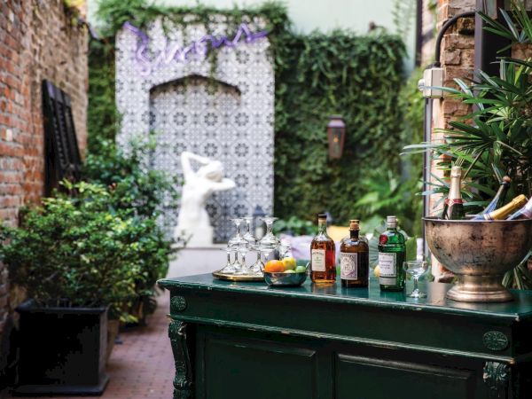 A cozy outdoor bar setup in a brick courtyard with drinks and utensils on a green counter, lush plants, and a statue in the background.
