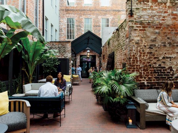 A cozy brick courtyard cafe with outdoor seating, tropical plants, and people chatting under a covered walkway.