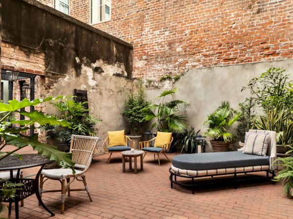 A small urban patio with brick floor, wicker chairs, lounge chair, and lush potted plants along high brick walls. End.