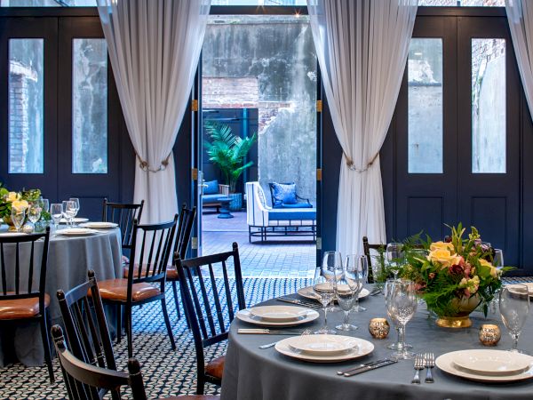 A stylish dining room with round tables, place settings, chairs, and an open doorway leading to a terrace with greenery.