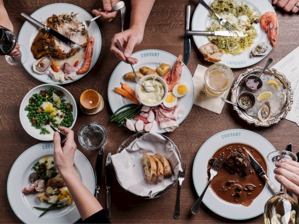 A table full of varied meals: plates with roasted meats, greens, sides, plus drinks and cutlery; several hands reach in to share the feast.