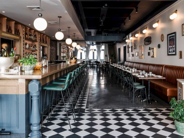 A stylish, vintage cafe interior with checkered floor, long row of tables and chairs, a bar on the left, warm lighting, and plants.