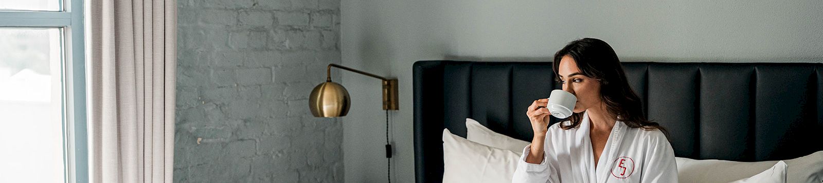 A woman in a white robe sits on a white bed, drinking from a cup in a bright, modern bedroom with a light gray brick wall and large window.