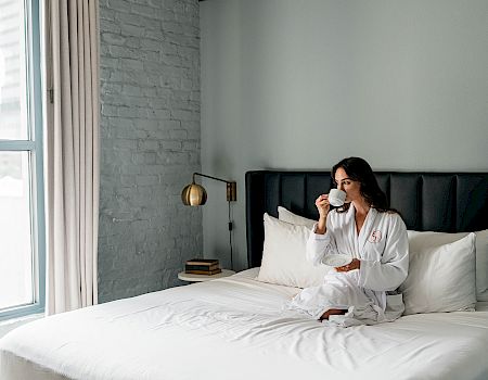 A woman in a white robe sits on a white bed, drinking from a cup in a bright, modern bedroom with a light gray brick wall and large window.