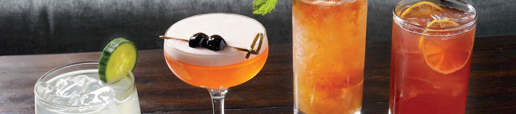 A row of four cocktails on a wooden bar: a pale lime drink with a lime wedge, a pink coupe with berries, and two tall, colorful drinks topped with mint and citrus.