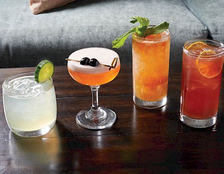 A row of four cocktails on a wooden bar: a pale lime drink with a lime wedge, a pink coupe with berries, and two tall, colorful drinks topped with mint and citrus.