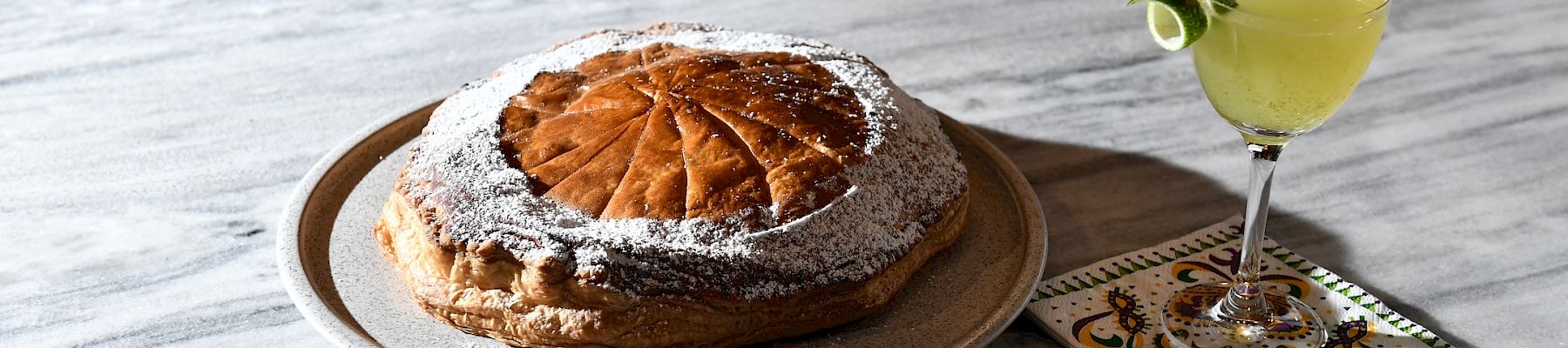 A round cake dusted with powdered sugar sits on a pale plate next to a small cocktail with a cucumber slice garnish on a marble table.