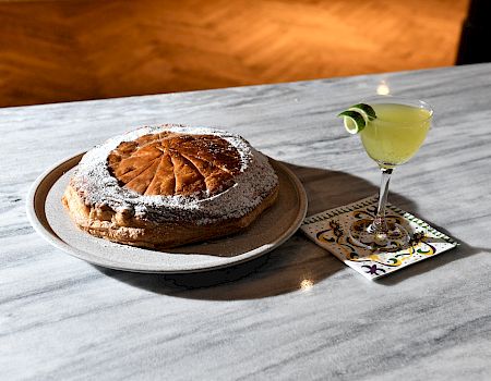 A round cake dusted with powdered sugar sits on a pale plate next to a small cocktail with a cucumber slice garnish on a marble table.