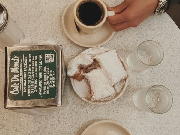 Two cups of coffee on a round table with a plate of sugar-dusted pastries, a green cigarette box, and a wooden-hand wearing a watch, shot from above.