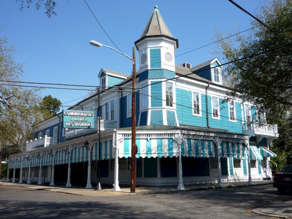 A bright turquoise, turreted two-story Victorian-style inn with a wraparound porch and striped awnings, sunny street scene.
