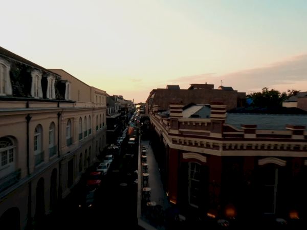 Sunset over a quaint city street flanked by classic buildings, narrow road lined with cars, soft orange glow casting long shadows.