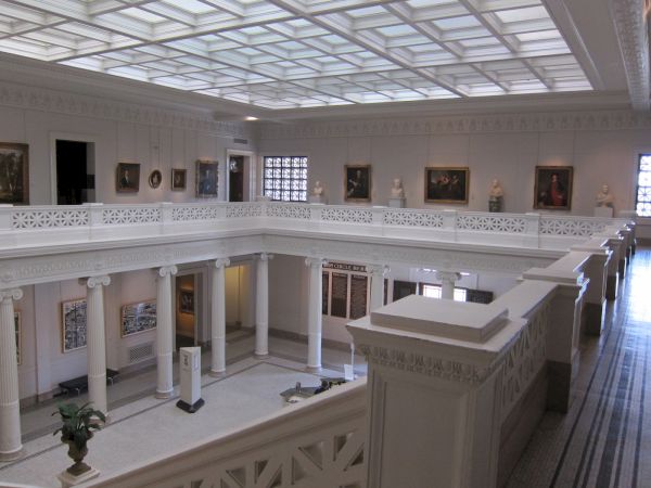 A grand, airy art museum interior with white pillars, open central atrium, balustrades, and framed paintings along the walls.