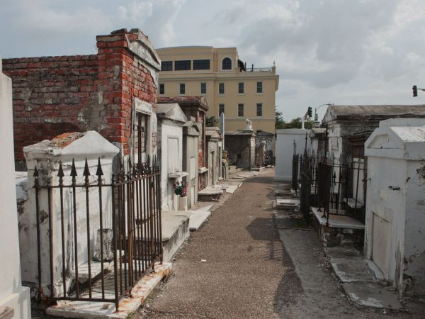 A narrow cemetery path lined with weathered mausoleums and iron fences on both sides, leading to a distant yellow building under a cloudy sky.
