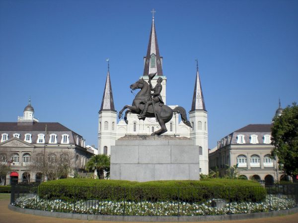 A statue of a rider on a horse sits atop a stone pedestal in a circular garden with white flowers, framed by a grand ch&acirc;teau-like building and blue sky.