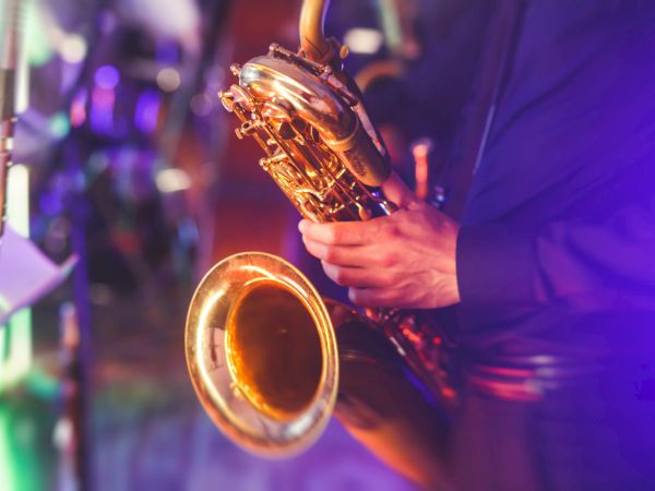 A close-up of a saxophonist playing a saxophone on a colorful, lit stage, with hands on the instrument and a microphone nearby, in a lively musical setting.