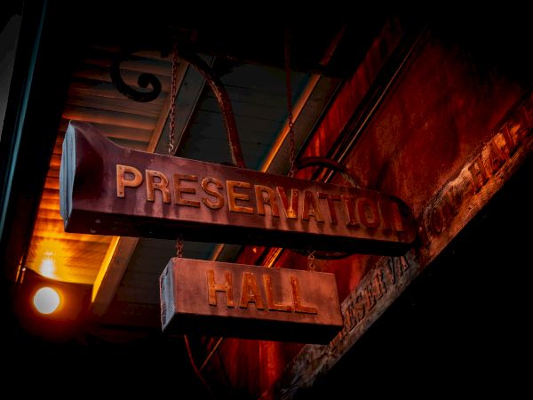 A dim, moody sign reads &ldquo;PRESERVATION HALL&rdquo; hanging from a weathered storefront, warm orange light casts eerie shadows.