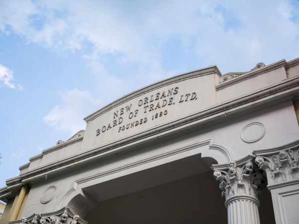 An ornate building facade with classical columns and a decorative plaque reading &ldquo;New Orleans Board of Trade, Ltd.&rdquo;, under a blue sky.