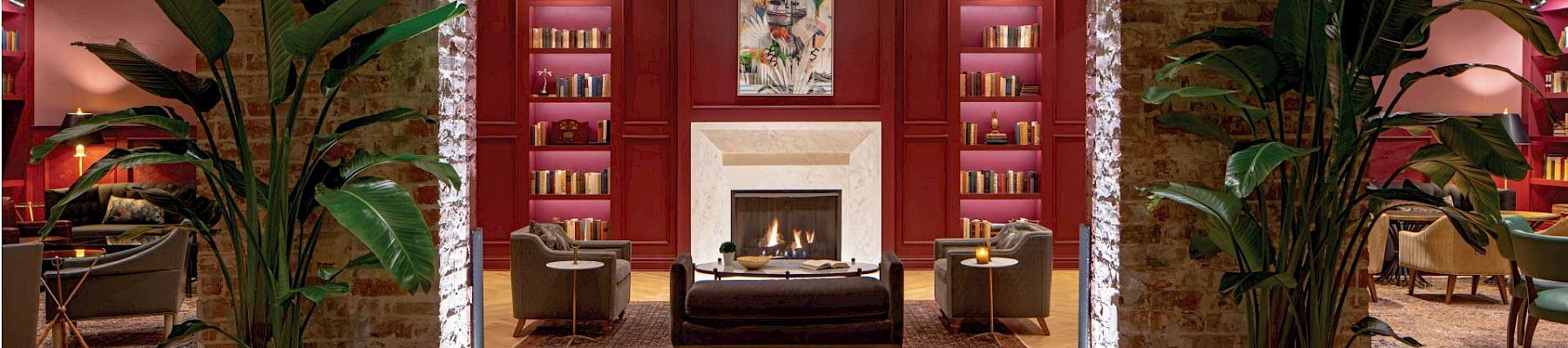 A stylish lobby or lounge area with a brick archway, fireplace, red walls, bookshelves, plants, and cozy seating&mdash;elegant and inviting.