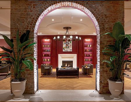 A stylish lobby or lounge area with a brick archway, fireplace, red walls, bookshelves, plants, and cozy seating&mdash;elegant and inviting.