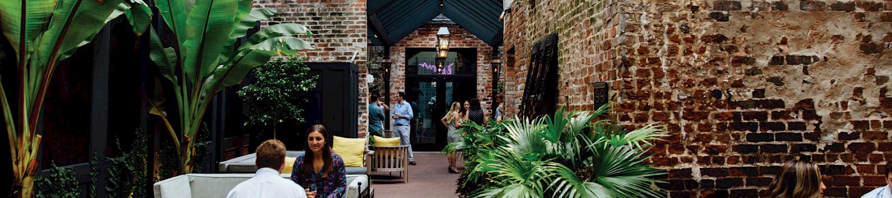 A cozy urban courtyard cafe with wicker seating, plants, and brick walls; people chat and sip drinks under a shaded entrance.