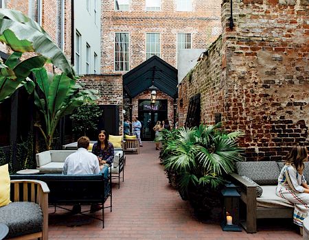 A cozy urban courtyard cafe with wicker seating, plants, and brick walls; people chat and sip drinks under a shaded entrance.